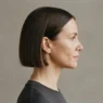 Woman with a chin-length blunt bob in side profile against a warm grey studio backdrop with soft front lighting