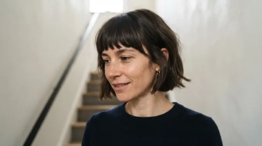 Woman with chin-length fringed bob standing on staircase, front fringe pieces visible as shorter face-framing sections in soft overhead light