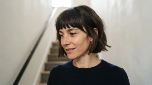 Woman with chin-length fringed bob standing on staircase, front fringe pieces visible as shorter face-framing sections in soft overhead light