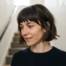 Woman with chin-length fringed bob standing on staircase, front fringe pieces visible as shorter face-framing sections in soft overhead light