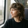 Woman with a modern medium length shag haircut in a train window seat, daylight across her hair, relaxed and natural
