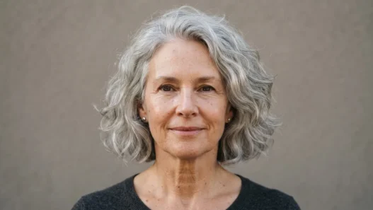 62-year-old woman with air-dried silver chin-length bob, soft daylight, warm grey backdrop, editorial framing from crown to collarbone