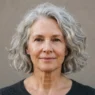 62-year-old woman with air-dried silver chin-length bob, soft daylight, warm grey backdrop, editorial framing from crown to collarbone