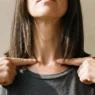 Woman pressing fingertips to collarbone to mark textured lob length reference point, hair hanging smooth