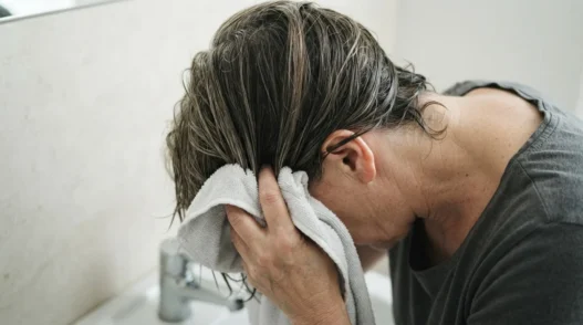 52-year-old woman squeezing microfibre towel upward into damp mid-length layered hair over bathroom sink