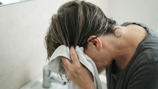 52-year-old woman squeezing microfibre towel upward into damp mid-length layered hair over bathroom sink