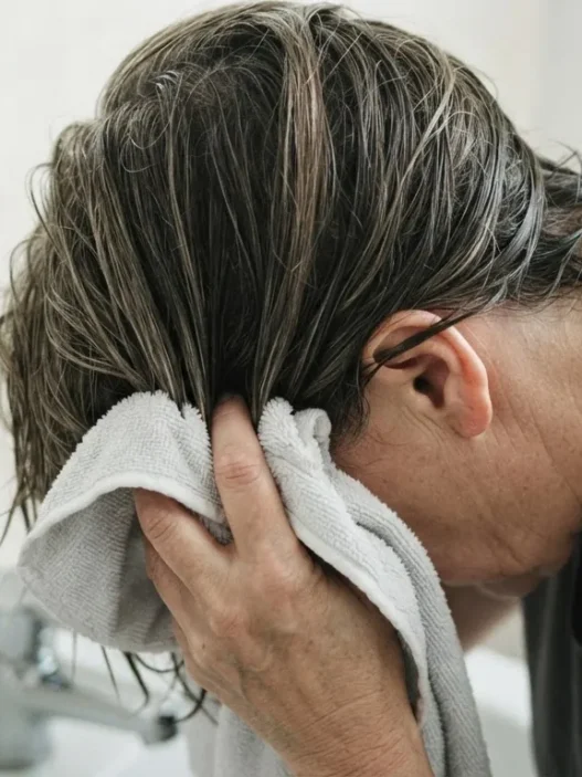 52-year-old woman squeezing microfibre towel upward into damp mid-length layered hair over bathroom sink