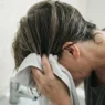 52-year-old woman squeezing microfibre towel upward into damp mid-length layered hair over bathroom sink