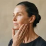 51-year-old woman with hair pulled back pressing her hand to her cheek assessing jaw and face shape in natural light
