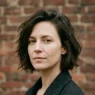 Woman with tousled chin-length bob in three-quarter view against brick wall in daylight