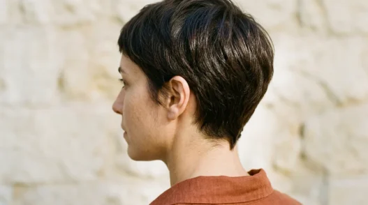 Woman with dark chestnut tapered pixie showing clean nape shape against stone wall