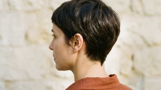 Woman with dark chestnut tapered pixie showing clean nape shape against stone wall
