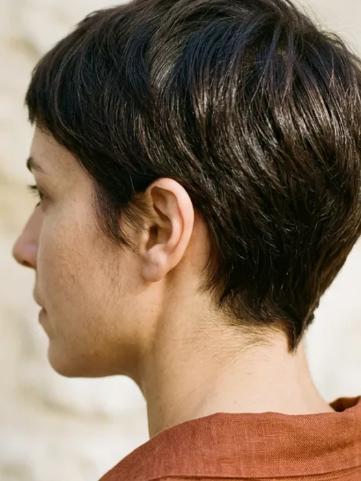 Woman with dark chestnut tapered pixie showing clean nape shape against stone wall